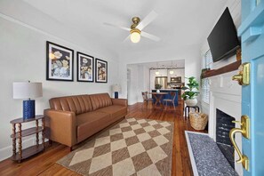 A living room with a brown leather sofa, a checkered rug, a ceiling fan, framed floral artwork, and a television mounted on the wall. - The Dellwood Cottage - Near Local Shops & Eats (Jacksonville)