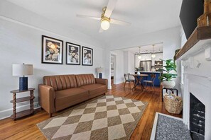 Un salon avec un canapé marron, un tapis à carreaux, un ventilateur de plafond et des œuvres d’art florales encadrées accrochées au mur.