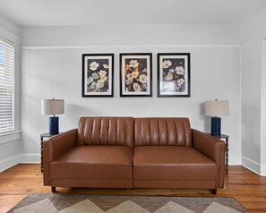 A brown leather sofa in a room with white walls and a patterned rug. There are three framed floral artworks on the wall. - The Dellwood Cottage - Near Local Shops & Eats (Jacksonville)