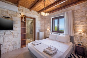 A bedroom with a stone wall, wooden ceiling, a bed with white linens, a TV, and a window with curtains. - Villa Freedom | A Private Luxury Estate in the Heart of Crete (Chania)