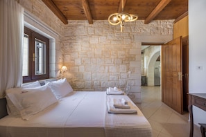 A bedroom with a stone wall, wooden ceiling beams, a bed with white linens, a bedside lamp, and a view into the kitchen. - Villa Freedom | A Private Luxury Estate in the Heart of Crete (Chania)