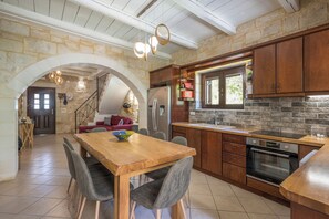A kitchen with a stone wall, wooden cabinets, a dining table with chairs, and a built-in oven. - Villa Freedom | A Private Luxury Estate in the Heart of Crete (Chania)