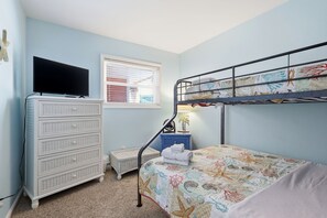 Un lit superposé avec une couvre-lit à motifs, un téléviseur posé sur une commode, une fenêtre avec des stores et une petite table de chevet avec une lampe.