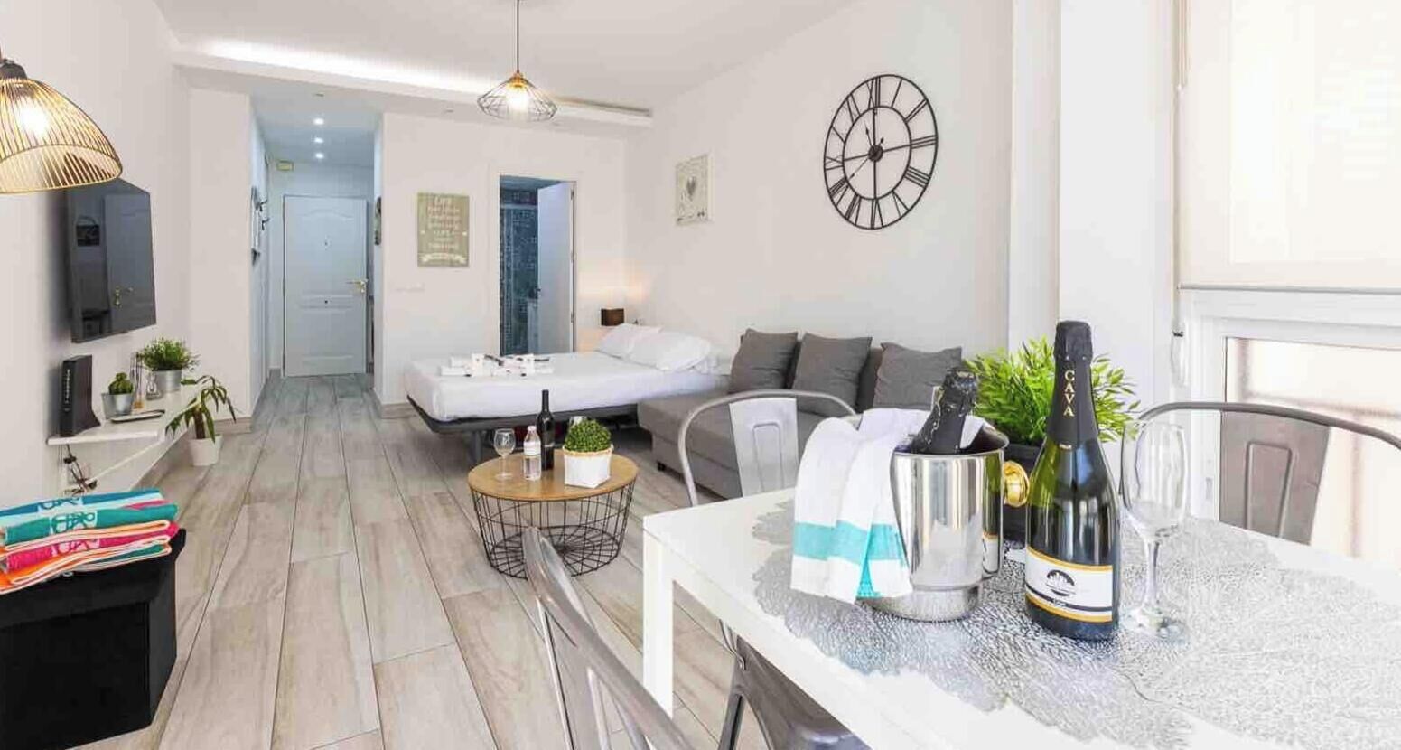 A modern living room with a sofa, a dining table with a bottle of champagne, and a wall clock.