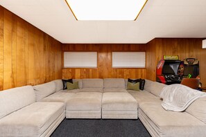 Ein Zimmer mit Ecksofa, zwei Fenstern mit Jalousien und einer Rennspielmaschine.