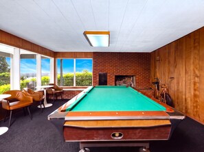 A pool table in a room with wooden walls and a brick fireplace. There are chairs and a table near the windows. - Bay View Dunes Getaway (North Bend)
