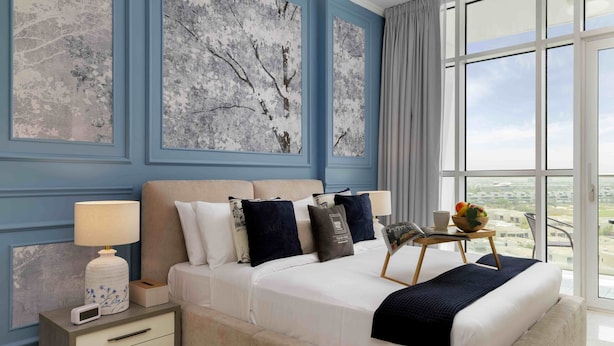 A hotel room with a large bed, a bedside table with a lamp, a small table with a fruit bowl, and a view of the city through large windows. - Elegant Studio Retreat in Damac Hills (Dubai)
