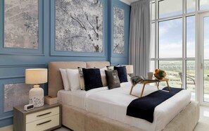 A hotel room with a large bed, a bedside table with a lamp, a small table with a fruit bowl, and a view of the city through large windows. - Elegant Studio Retreat in Damac Hills (Dubai)