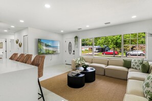 A modern living room with a white sofa, a dining area with wicker chairs, a large window with a view of the street, and a flat-screen TV mounted on the wall. - Tampa Pool Oasis, Lux Boho Design, , Otdr Movie & Kitchen, Near Golf (Tampa)