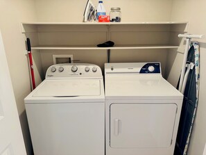 A laundry room with two front-loading washing machines, a shelf above with cleaning supplies, and a door with a towel rack. - Hawks Nest | 2BR, Ground, 3miles to Sports Complex + Free Perks (Mesquite)