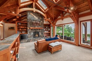 A spacious living room with a stone fireplace, wooden bookshelves, a leather sofa, and large windows offering a view of the outdoors. - Bottle Bay Lakefront Lodge | Dock & Game Room (Sagle)