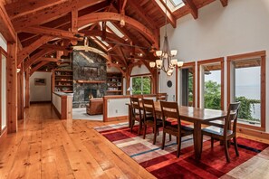 A spacious dining area with a large wooden table and chairs, a stone fireplace, and a view of the outdoors through large windows.