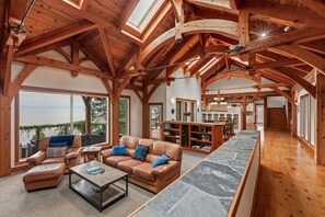 A spacious living area with a high ceiling, wooden beams, a stone fireplace, leather sofas, and a long dining table. - Bottle Bay Lakefront Lodge | Dock & Game Room (Sagle)
