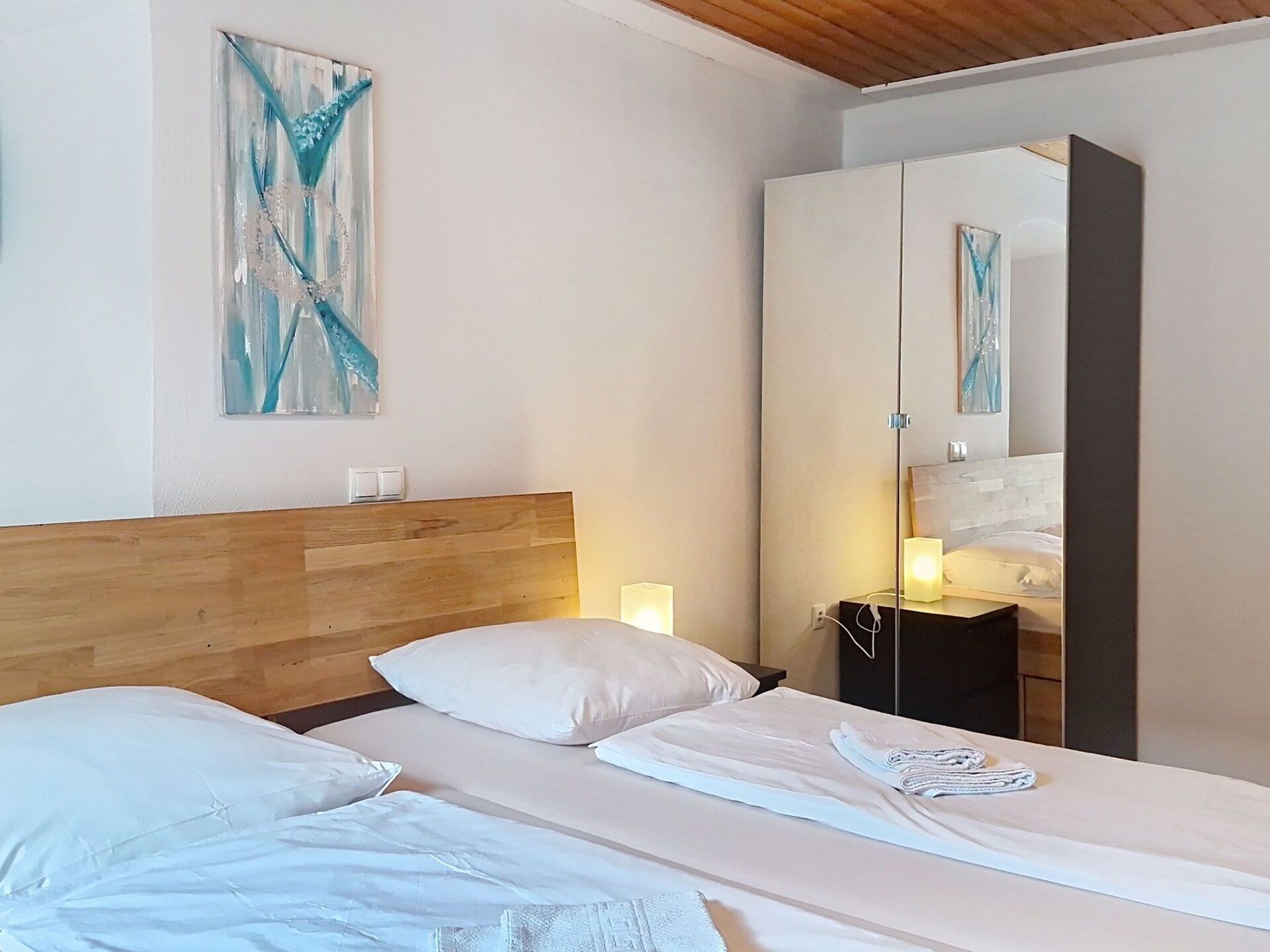 A hotel room with a wooden headboard, a bed with white linens, a bedside table with a lamp, and a large mirror reflecting the room.