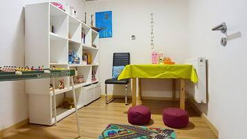 A room with a white bookshelf, a small table with a yellow tablecloth, two purple stools, and a colorful rug on the floor.