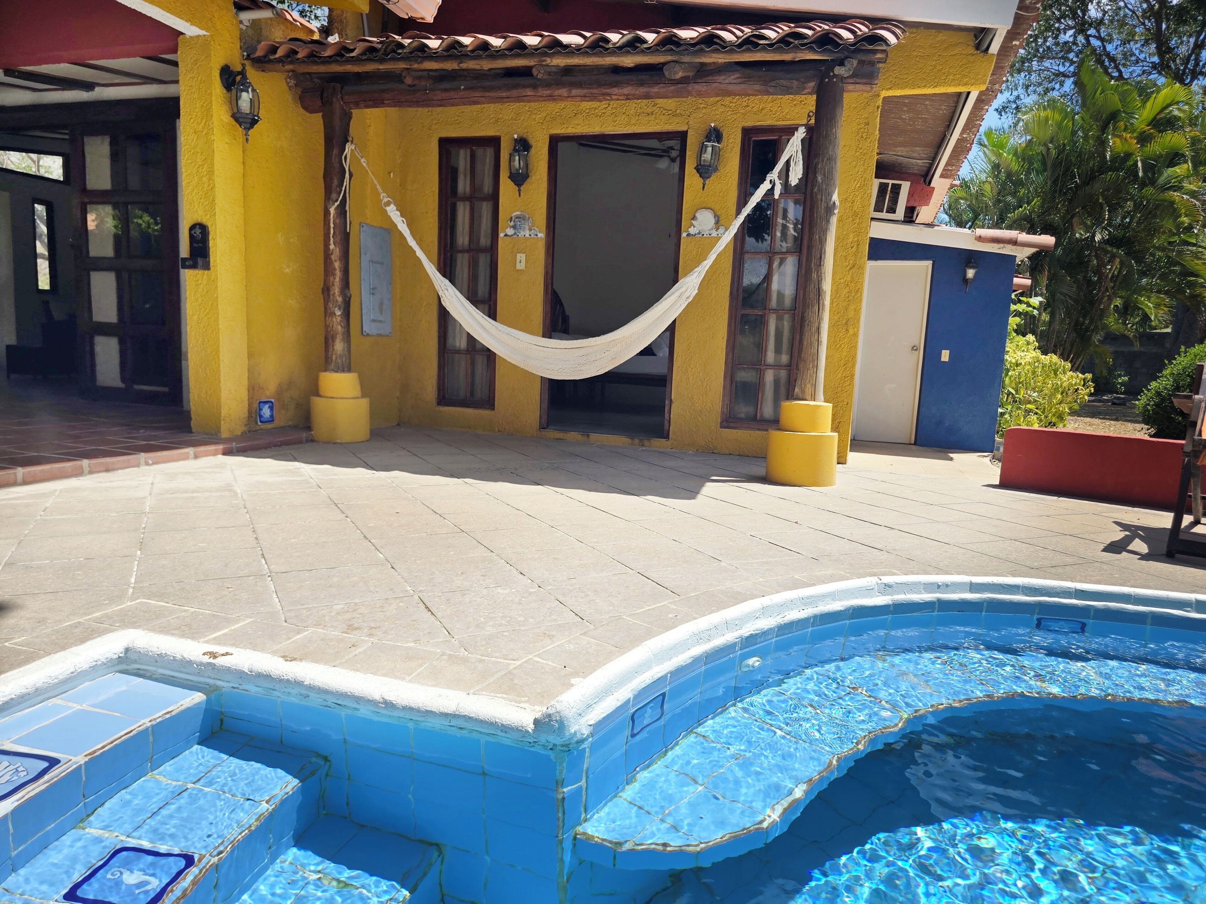A pool area with a hammock, a yellow building, and a blue pool.