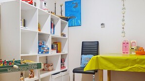 Une pièce avec une bibliothèque blanche, un bureau recouvert d’une nappe jaune, une chaise et un meuble rose.