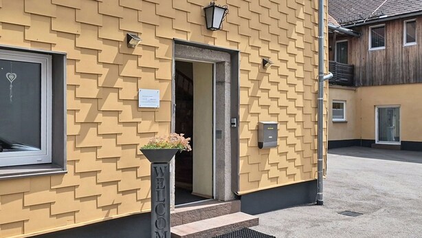 Un bâtiment à la façade en briques jaunes, une porte ornée d’une fenêtre en forme de cœur et un jardinier avec des fleurs.