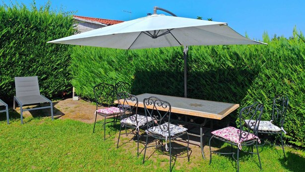 Um pátio externo com mesa e cadeiras, um grande guarda-sol e um hedge ao fundo.