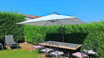 Eine Außenpatio mit Tisch und Stühlen, einem großen Sonnenschirm und einer Hecke im Hintergrund.