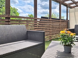 Un espacio al aire libre con una pérgola de madera, sofás grises trenzados y una planta en maceta con flores amarillas.