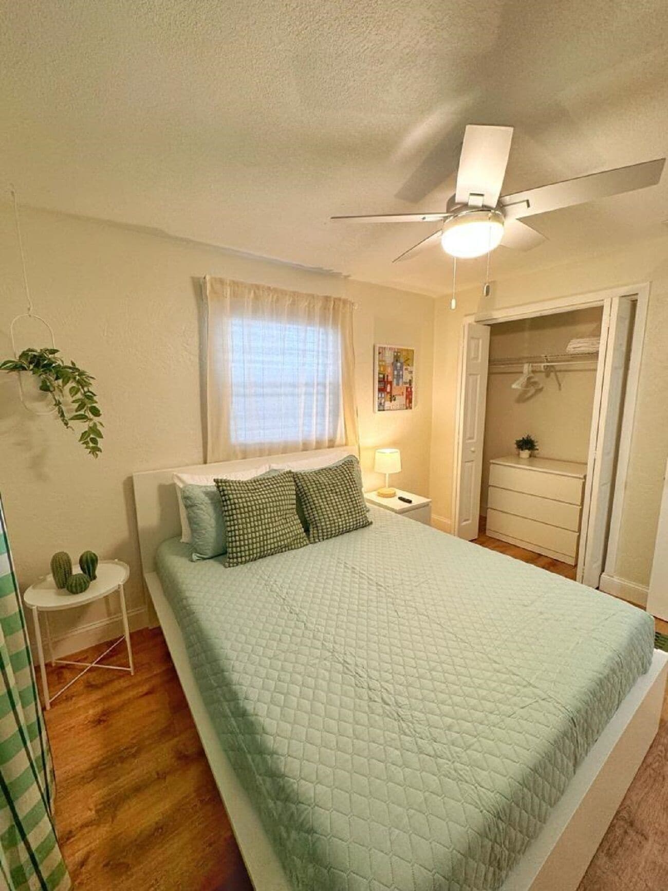Ein Schlafzimmer mit einem Bett, einem Deckenventilator, einem Fenster mit Vorhängen, einem Nachttisch mit Lampe und einer Kommode.