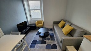 Un salon moderne avec un canapé, des coussins jaunes, une table basse ronde, un téléviseur à écran plat et un tapis à motifs.