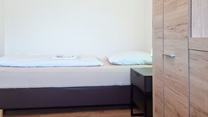 Una cama individual con colchón gris y ropa de cama blanca, una mesita de noche pequeña y un armario de madera.