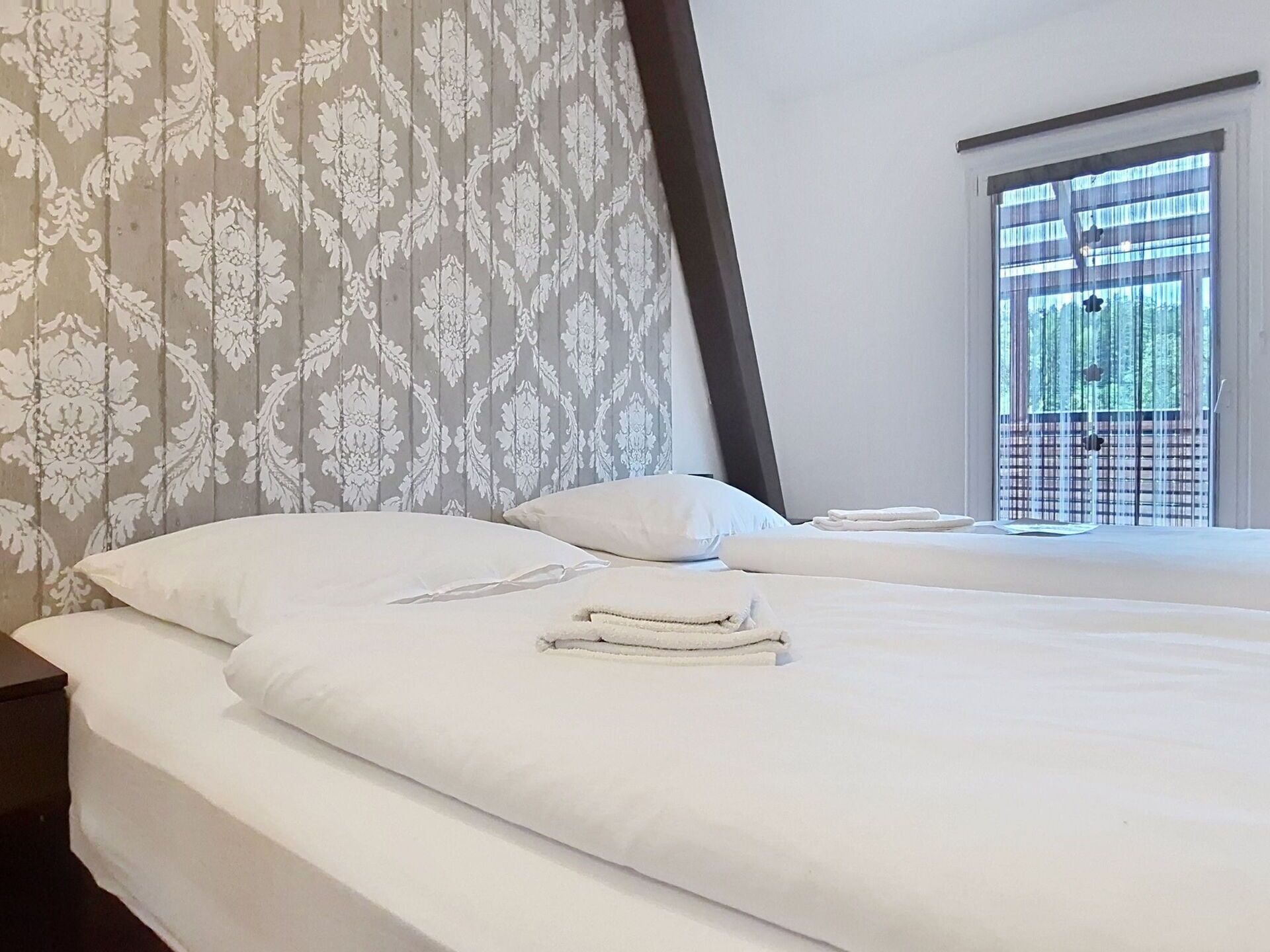 A hotel room with two beds, white linens, a patterned wallpaper, and a window with a view of trees.