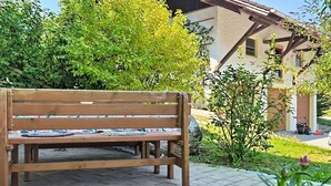 Un banc en bois avec un coussin, entouré de verdure et bordé d’une zone pavée.