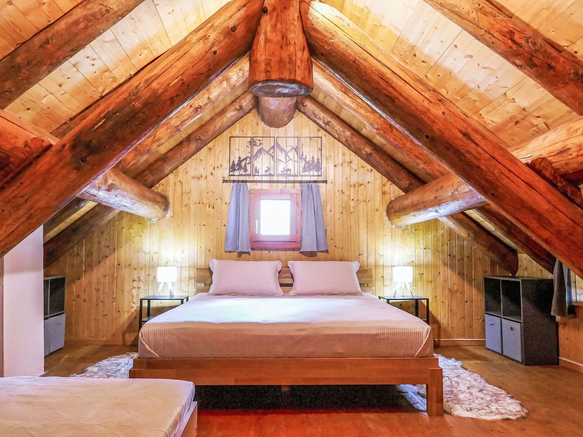 A cozy attic bedroom with a wooden sloped ceiling, a large bed, two bedside tables, and a small window with curtains.