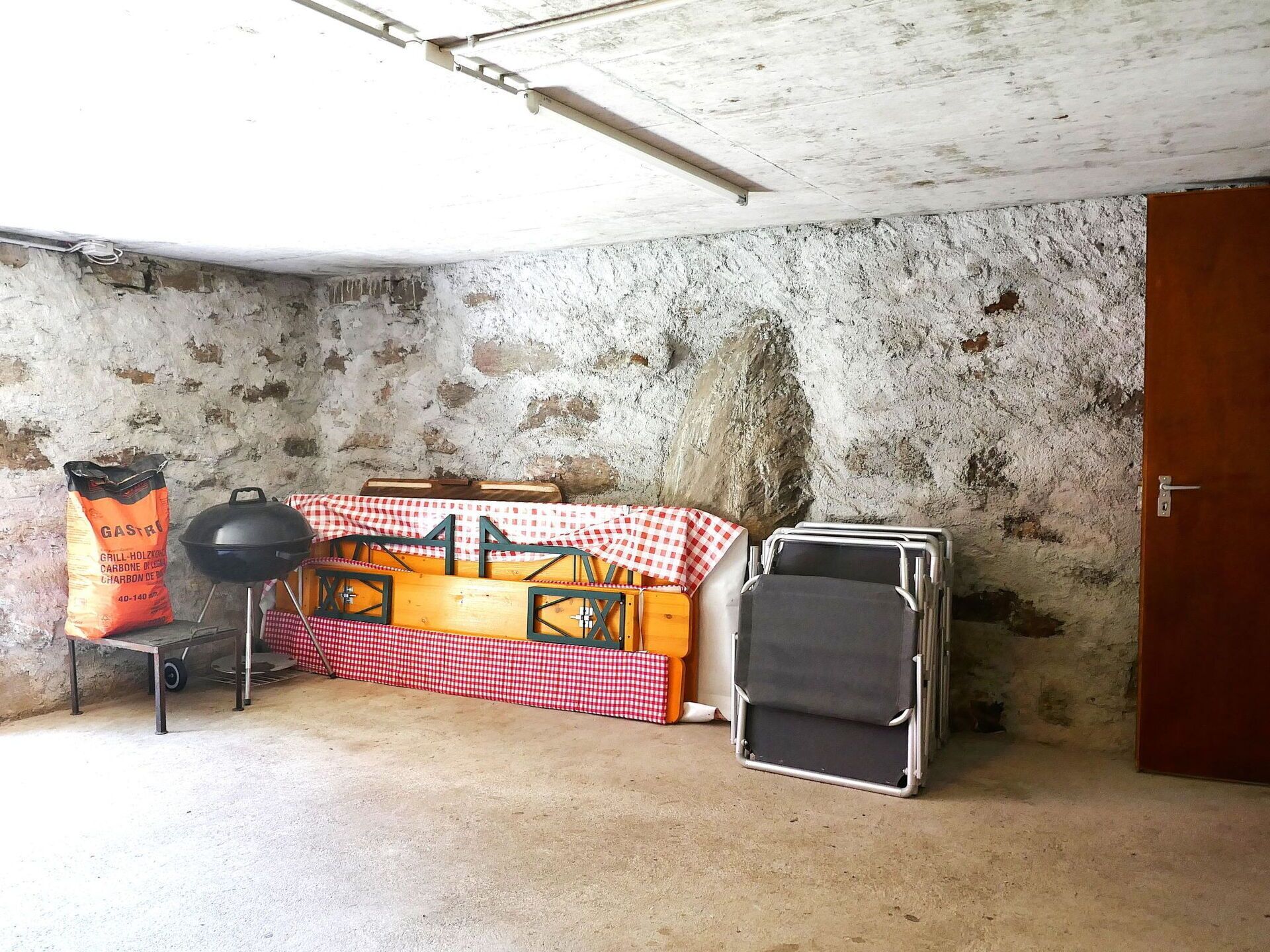A room with a concrete wall, a wooden bench with a checkered tablecloth, a grill, and a suitcase.