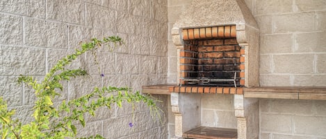 Un four extérieur en pierre, avec un intérieur en briques, une étagère en bois et une plante en pot placée à côté.
