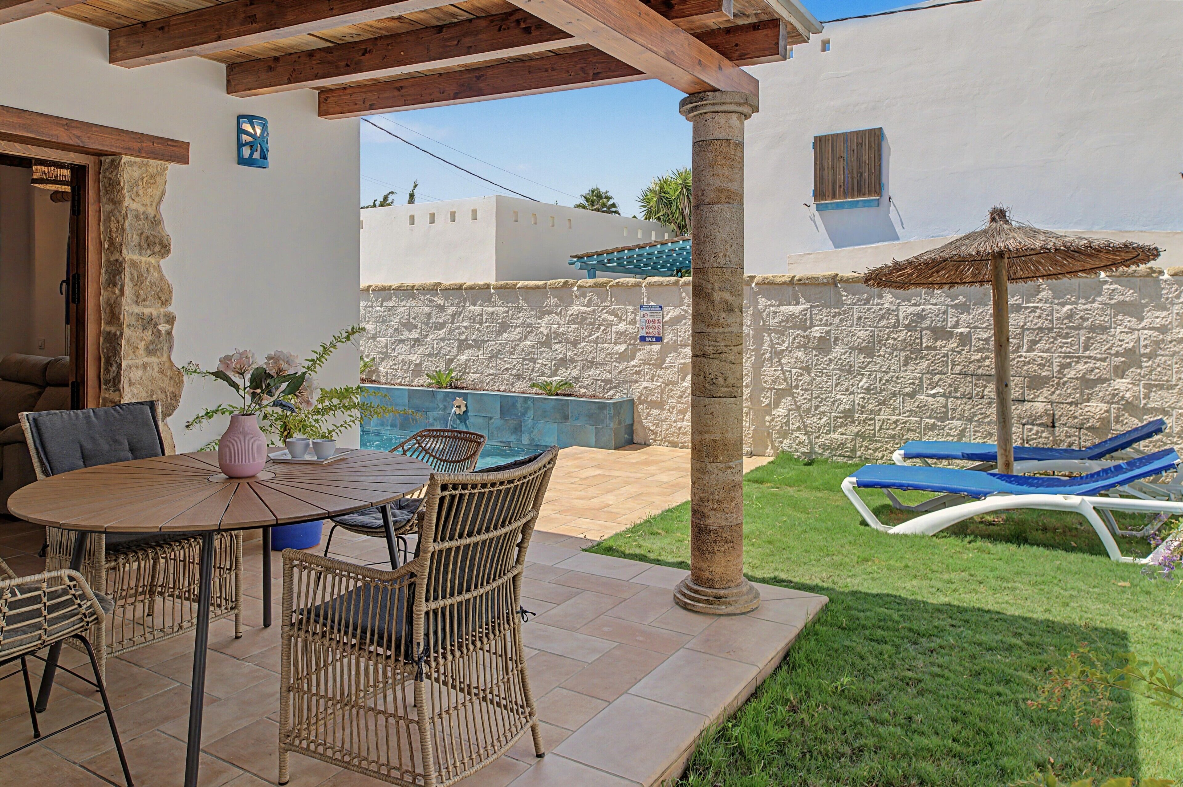 A patio area with a round table and wicker chairs, a stone wall, a thatched umbrella, and blue lounge chairs.