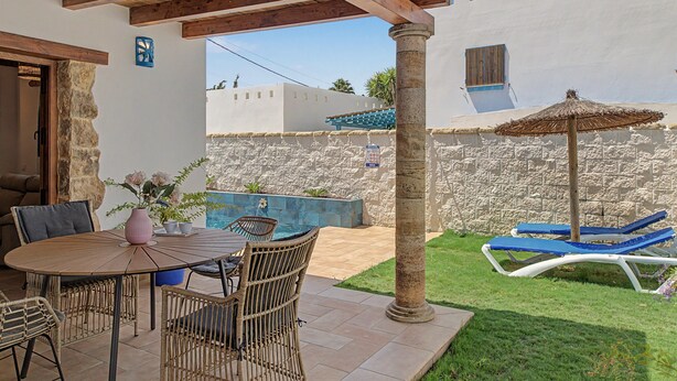 Un patio con una mesa redonda y sillas de mimbre, un muro de piedra, un paraguas de paja y sillones azules.