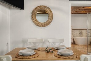 Ein Essbereich mit einem Holztisch, vier weißen Tellern, zwei weißen Stühlen und einem gewebten Spiegel an der Wand.