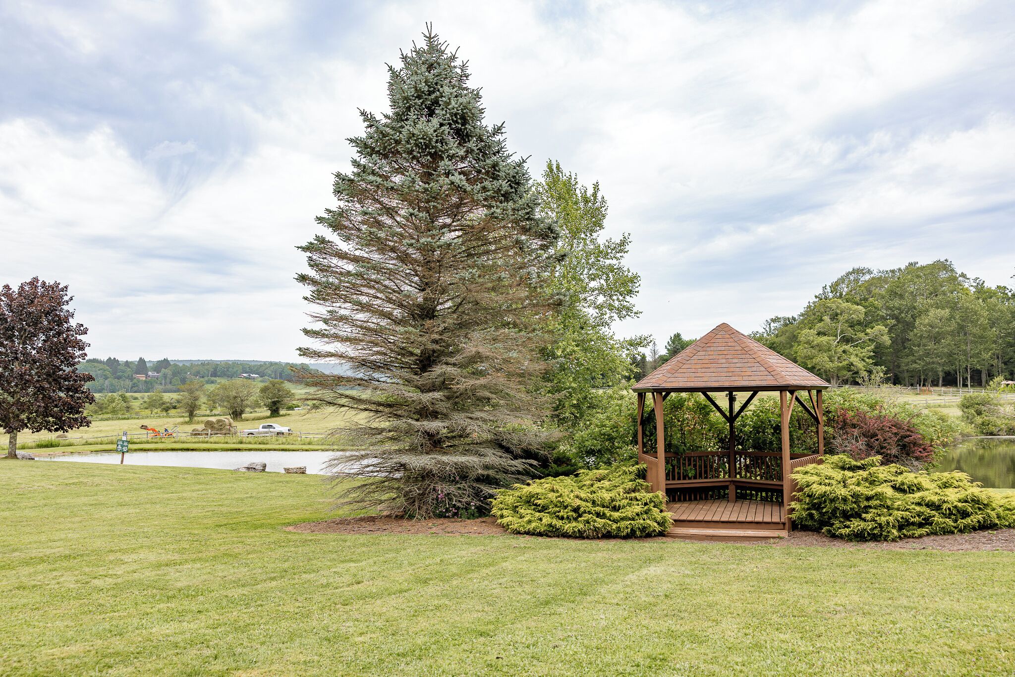 Un belvédère doté d’un toit en tuiles, entouré de pelouse et d’arbustes bien entretenus, situé près d’une étendue d’eau et d’un grand arbre à feuilles persistantes.