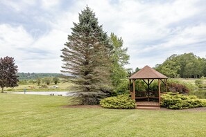 A gazebo with a tiled roof, surrounded by well-maintained grass and shrubs, near a body of water and a large evergreen tree.