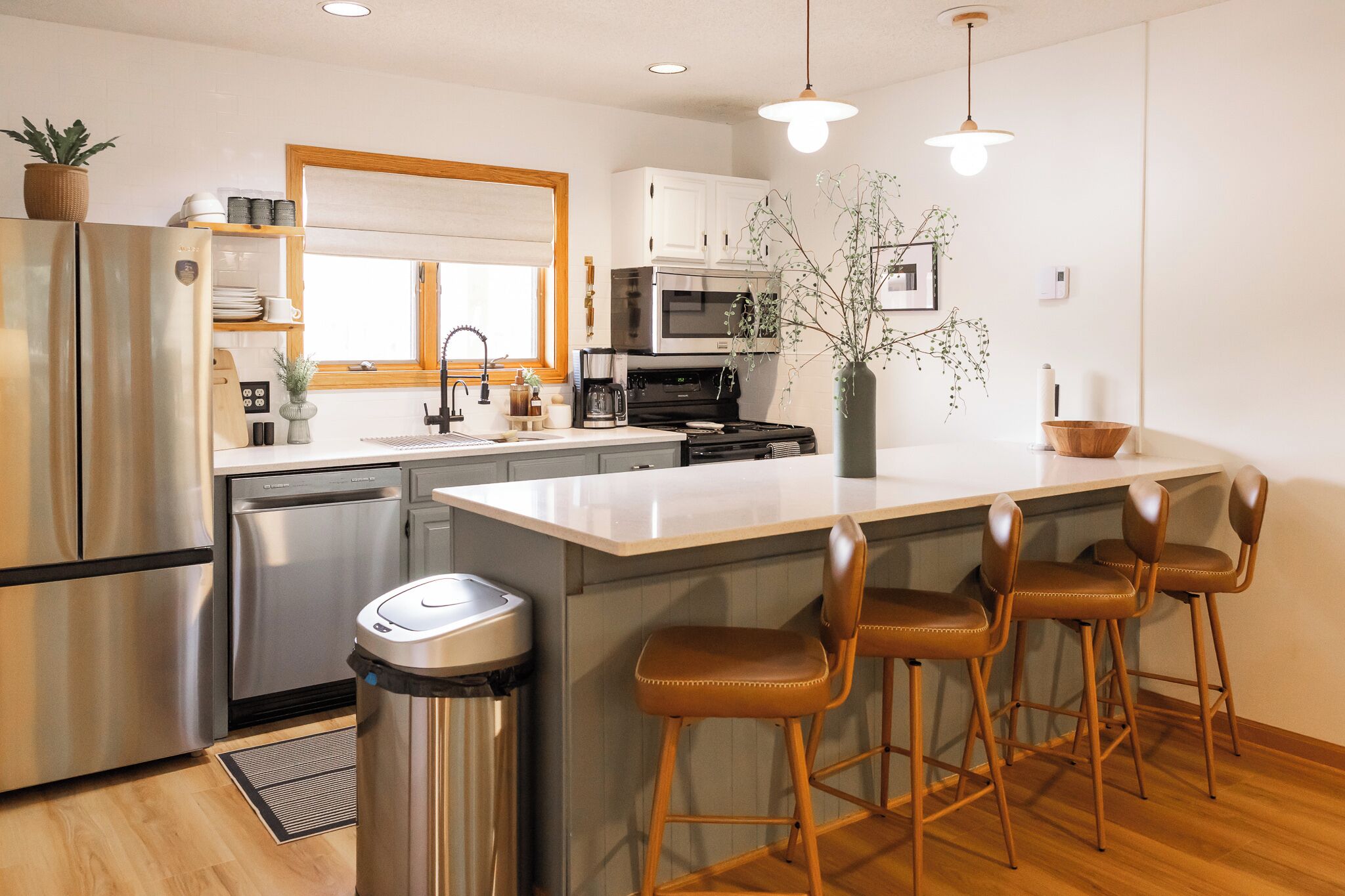 A modern kitchen with a stainless steel refrigerator, a built-in oven, a microwave, and a sink with a wooden countertop. There are three bar stools and a trash can.