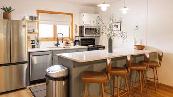 A modern kitchen with a stainless steel refrigerator, a built-in oven, a microwave, and a sink with a wooden countertop. There are three bar stools and a trash can.