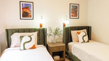 A hotel room with two beds, each with a green headboard, white bedding, and decorative pillows. There are two wall-mounted lamps and a small table with a vase between the beds.