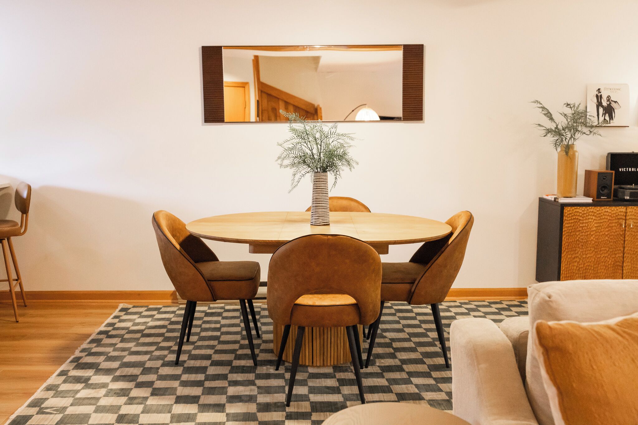 Un coin repas avec une table ronde, quatre chaises, un vase avec des plantes, un miroir au mur et un tapis à carreaux.