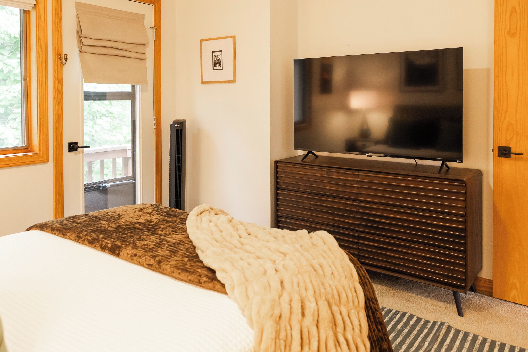 A bedroom with a bed, a television on a wooden dresser, a window with a view, and a door.