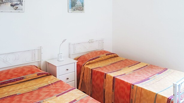Quarto simples e limpo com duas camas de solteiro, uma mesa de cabeceira e um criado-mudo.