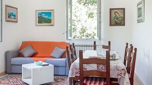 Uma sala de estar com sofá, mesa de jantar com cadeiras, janela com vista para vegetação e quadros nas paredes.