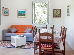 Ein Wohnzimmer mit Sofa, einem Esstisch mit Stühlen, einem Fenster mit Blick auf Grünflächen und gerahmten Bildern an den Wänden.