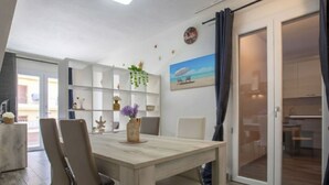 Uma área de refeições com mesa e cadeiras de madeira, uma estante branca com objetos decorativos e vista para o exterior por uma porta de vidro.
