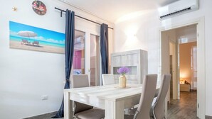 Uma sala de jantar com uma mesa de madeira, quatro cadeiras, um vaso com flores, um relógio de parede e uma pintura de uma praia.