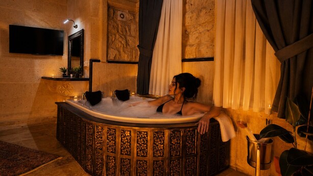 A woman relaxing in a freestanding bathtub with intricate woodwork, a stone wall, and a mounted television. - Uçhisar Palace in Cappadocia Hotel (Nevsehir)