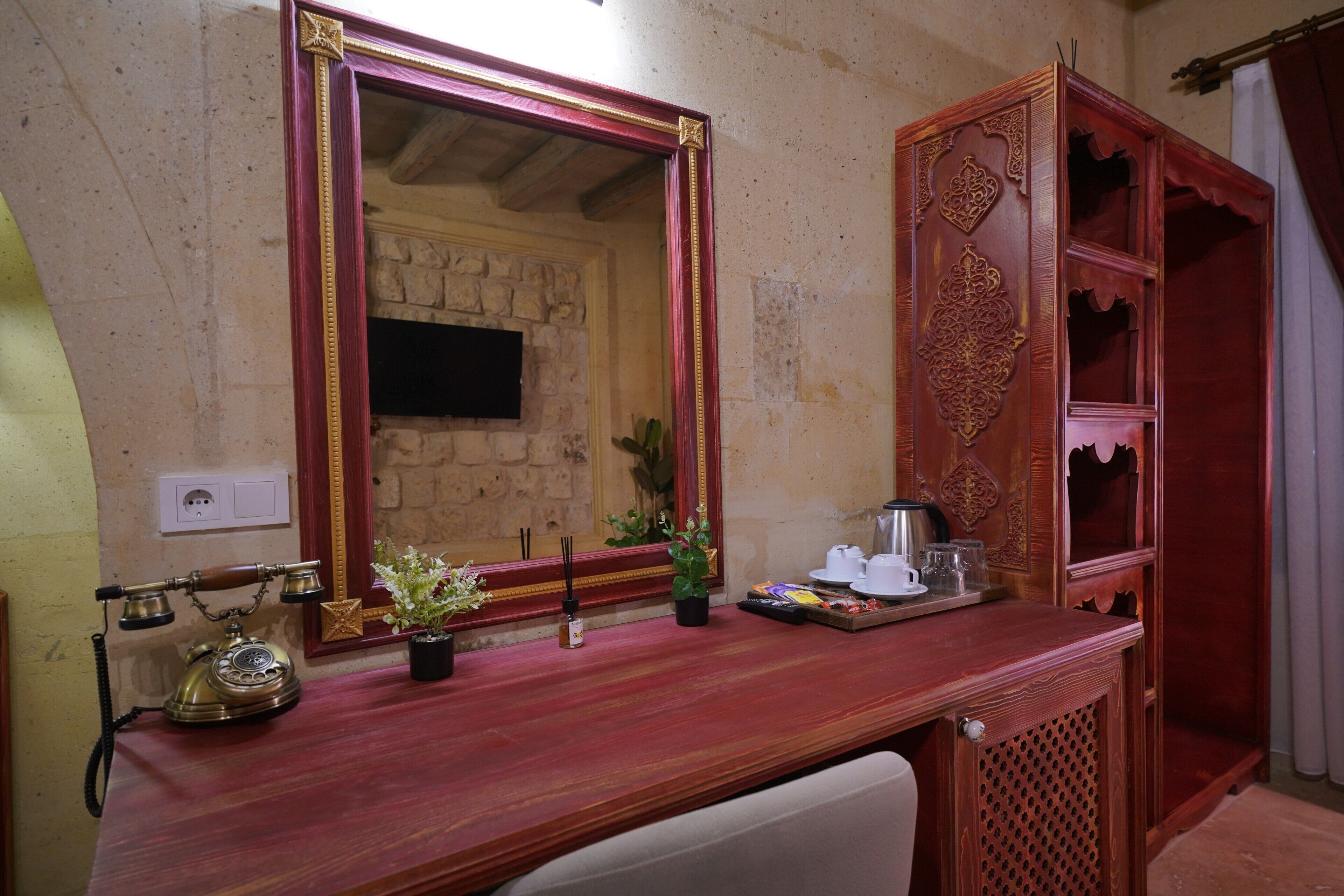 Una habitación con un escritorio de madera, un espejo, un teléfono, una planta pequeña y un armario con detalles decorativos tallados.